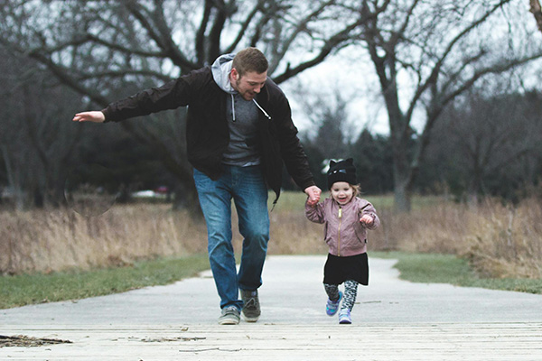 Dad and young toddler daughter outside walking down a path