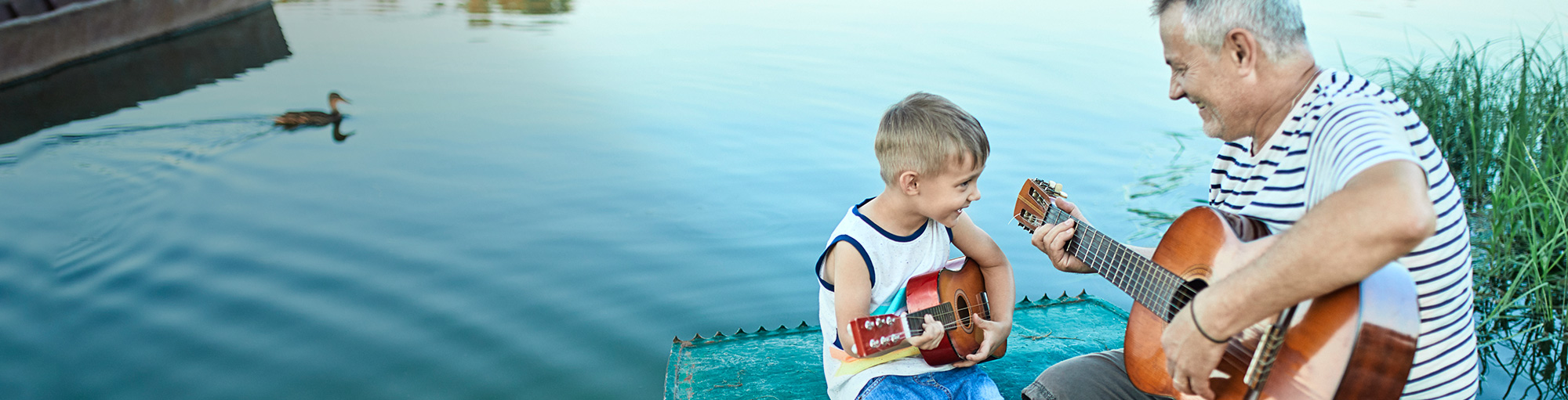 Grandfather playing guitar with grandson playing ukulele on a boat at the edge of a small pond